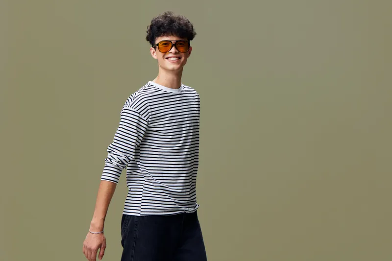 Smiling young man in striped shirt and sunglasses standing against plain background, casual style, positive mood, warm lighting, fashion portrait.