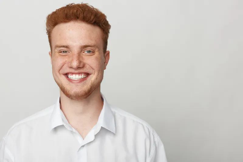 Smiling young man with red hair and beard wears white shirt, looking at camera on light grey background. Concept of happiness, confidence, and casual lifestyle portrait.