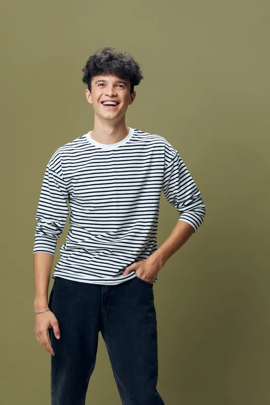smiling young man with curly hair wearing striped shirt and black pants standing against olive background in casual pose, happy expression