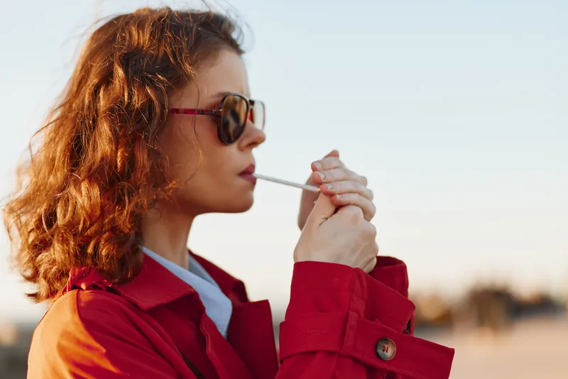Young woman with curly hair wearing a red coat lights a cigarette outdoors during golden hour, showing a calm and focused expression in a natural setting.