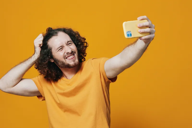 Happy man with curly hair taking a selfie against a vibrant yellow background, wearing a casual orange t shirt and expressing joy and confidence