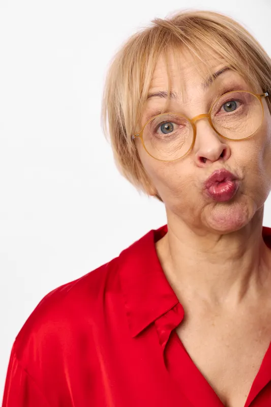 Surprised elderly woman with glasses, studio portrait on white background, red shirt, short blonde hair, wide open eyes and playful expression, closeup, soft light