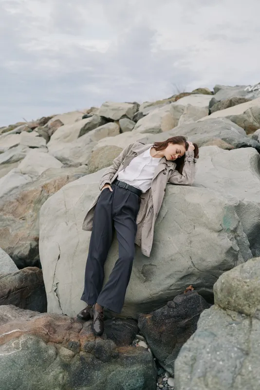 Young woman in coat and trousers leans against large rock on rocky beach under cloudy sky. Casual fashion portrait with natural landscape background and relaxed mood in outdoor setting.