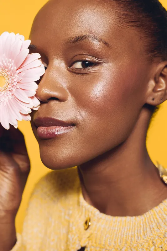 Close-up of a smiling black woman with dark skin holding a pink flower near her face. Yellow background and soft lighting emphasize natural beauty and skincare concept.