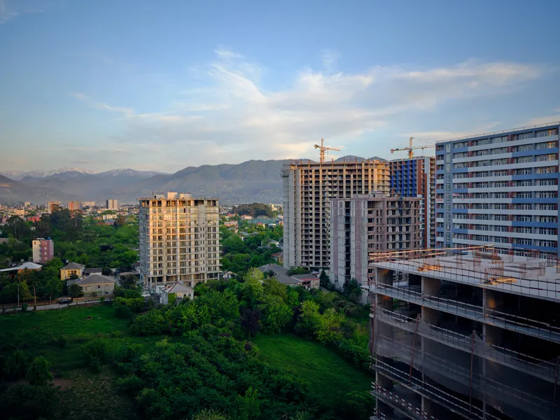 Construction buildings and cranes on skyline with apartment towers and city development, architecture details visible at sunset over green foreground and distant mountains.