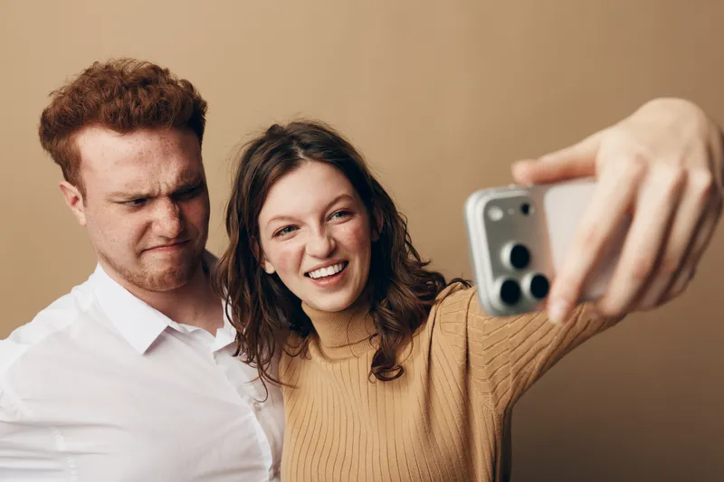Young man and woman take a selfie together, woman smiling and man making a funny face. Casual clothing and neutral background create a relaxed, playful mood for friendship concept.