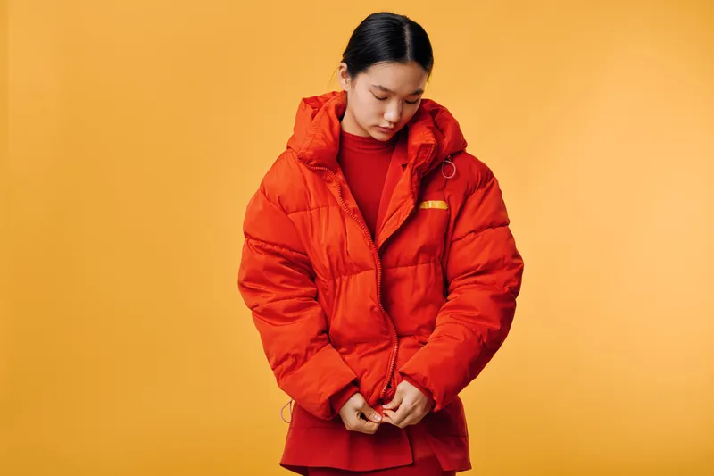 Woman wearing bright red warm jacket and sweater buttoning coat with thoughtful expression on yellow background indoors. Fashion, casual style and winter clothing concept.
