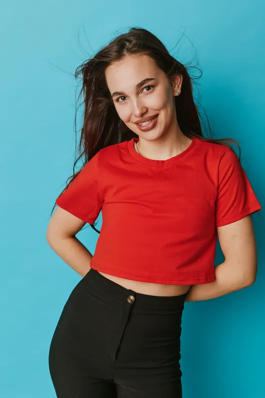 Young woman with long dark hair wearing red crop top and black pants posing with hands behind back smiling against blue background in studio portrait.
