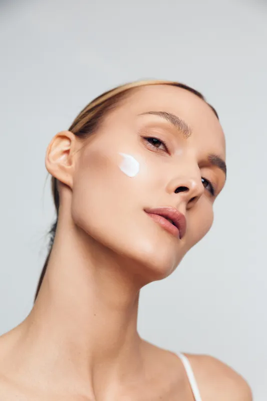 Young woman with clear skin applies moisturizing cream on cheek looks at camera with natural expression isolated on light background in skincare and beauty concept.