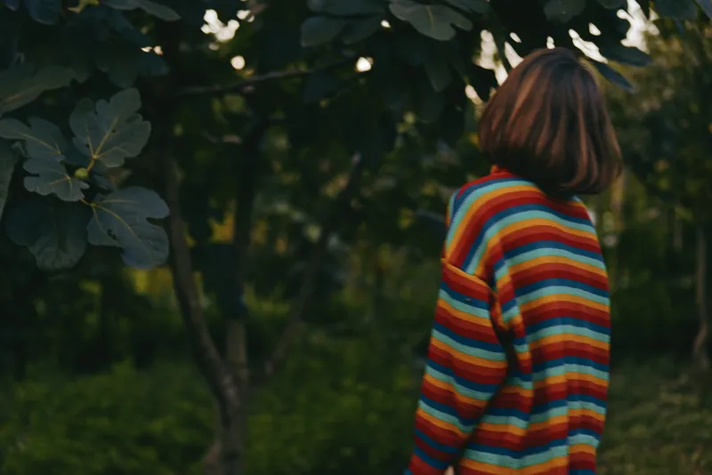 Woman in colorful striped sweater shown in back view with bob haircut, standing in nature among trees, contemplative mood, casual outfit for lifestyle travel and quiet outdoor exploration.