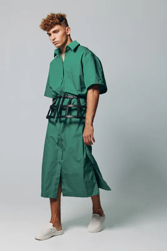 Young man posing in green long shirt dress with short sleeves and black style belt with perforations on neutral grey background inside studio space.