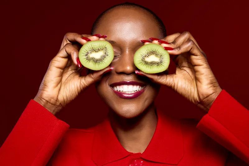 Smiling black woman with dark skin holds two kiwi halves in front of her eyes. She wears a red sweater and red nail polish, posing on a red background. Concept of healthy lifestyle and joy.
