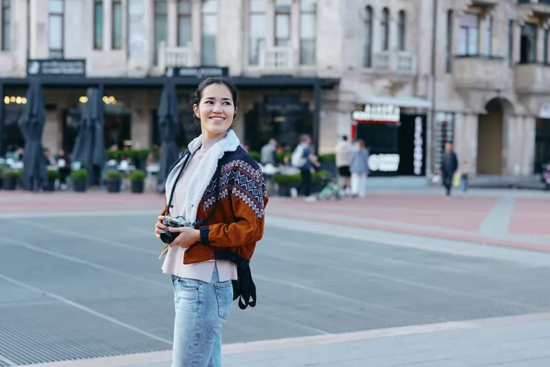 Woman with camera in a street plaza, casual chic jacket and scarf, smiling amid city backdrop, capturing candid moments in an urban daylight scene