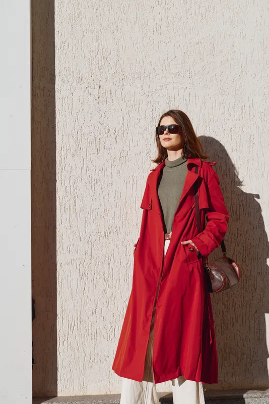 A stylish brunette woman wearing a bright red trench coat over an olive green top stands against a sunlit wall, exuding confident street fashion and bold urban attitude.