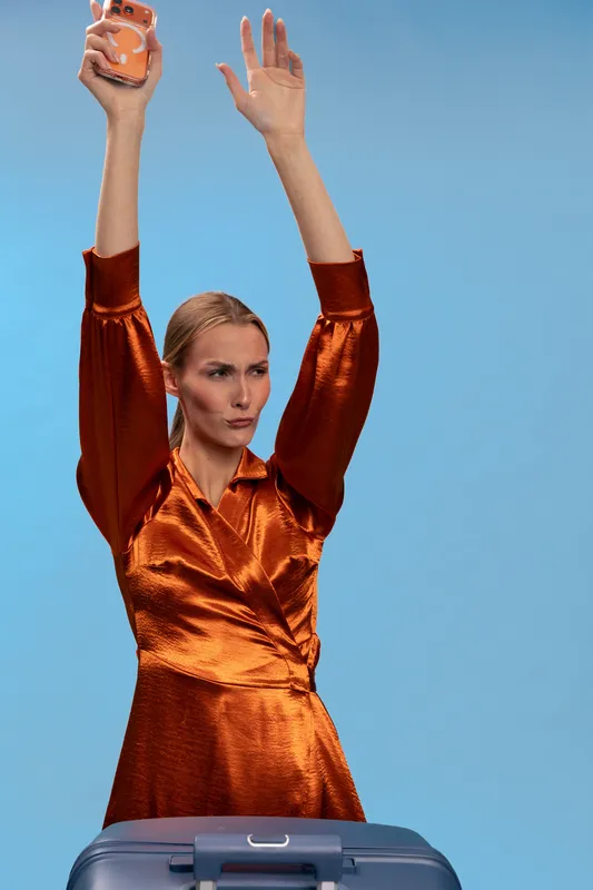 Confident woman in shiny orange dress holds phone above head with raised arms, standing behind suitcase on blue background. Concept for travel, celebration, success, and freedom.