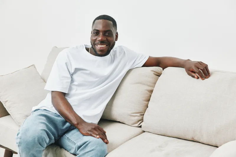 Smiling African American man relaxing on a beige couch in a casual white t shirt, embodying comfort and happiness at home Ideal for lifestyle and wellness themes