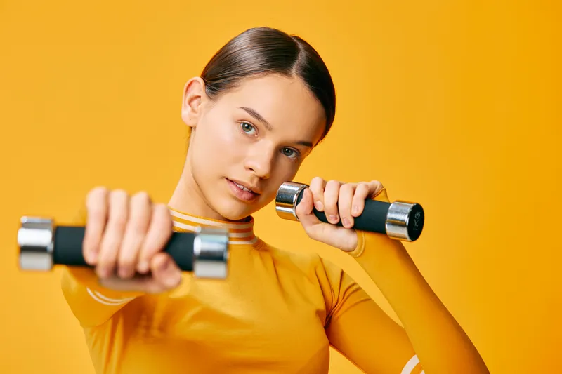 Young woman fitness portrait holding dumbbells in a focused workout pose, wearing yellow athletic top and matching sleeves against a solid orange studio background with clean composition.