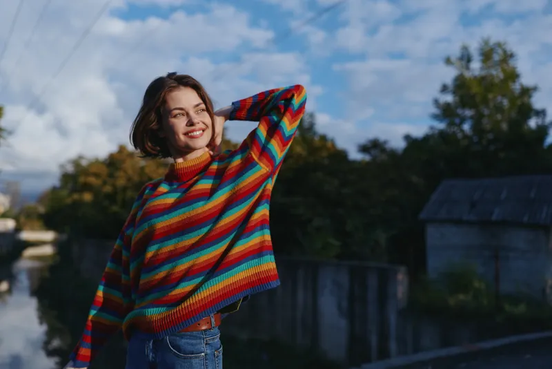 Young woman in rainbow sweater smiling outdoors on a suburban riverbank, casual outfit with denim jeans, leaning with hand on head, autumn travel lifestyle portrait in natural light.