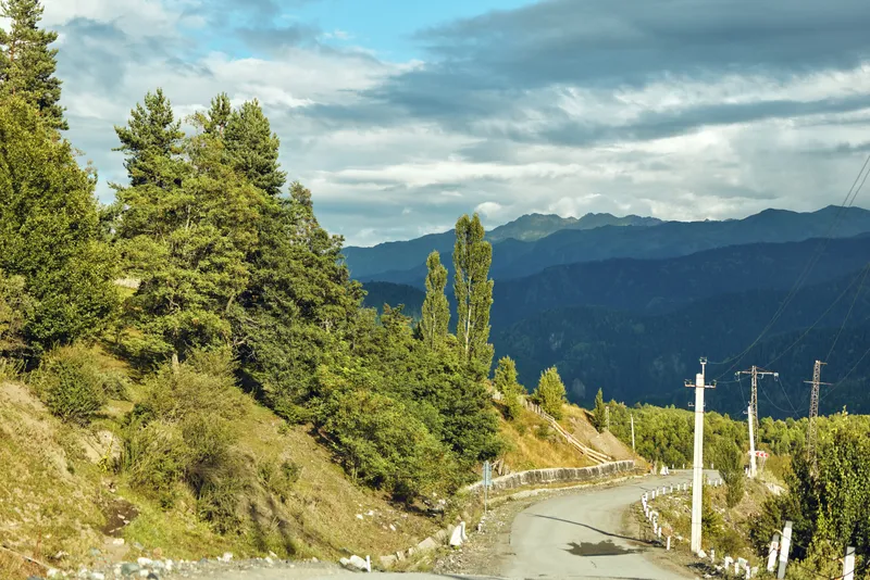 Mountain road winds through rugged landscape with trees and hills under a dramatic sky, offering a scenic countryside view ideal for travel and nature themed projects