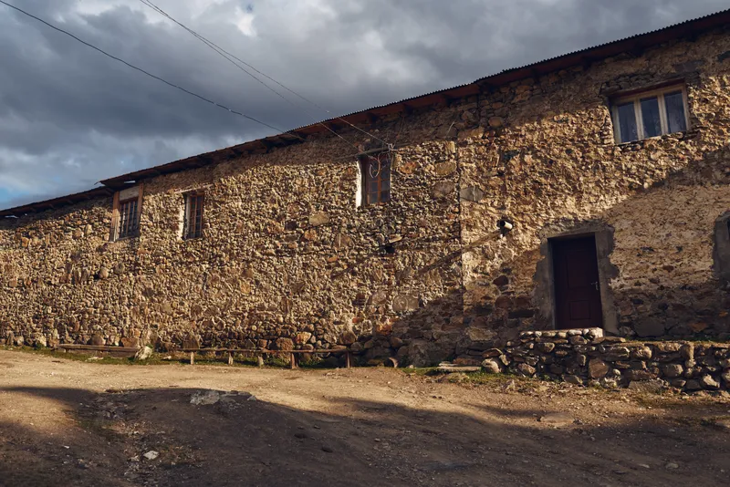 Weathered stone building in a rural setting with a dirt road and rocky ground, displaying a worn facade, small windows, and a simple doorway beneath a cloudy sky
