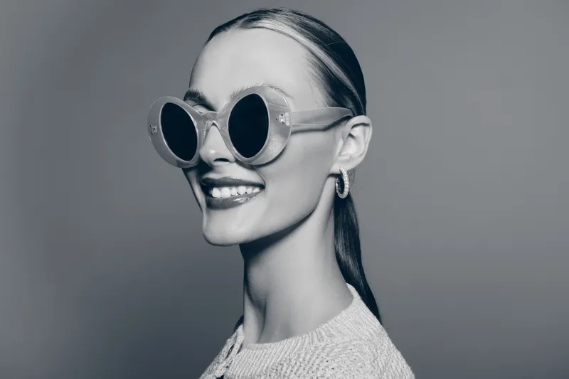 Smiling white woman with pale skin and slicked-back hair wears round sunglasses and hoop earrings. Stylish portrait in black and white studio with modern fashion concept.