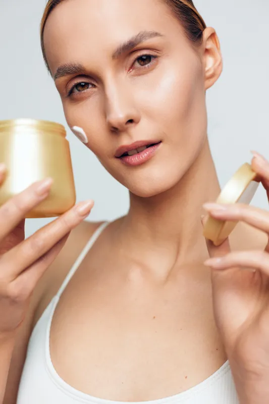 Close-up of a woman applying white face cream from a golden jar on her cheek. Skincare routine concept with soft lighting and natural skin texture visible on a plain background.