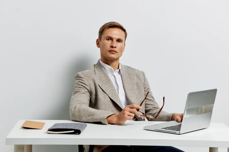 Professional man in beige blazer sitting at desk with laptop and glasses, looking serious and focused, workspace environment, business attire, modern office setting, indoors, neutral background, front