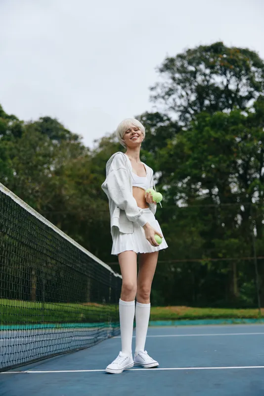 Young female athlete smiling joyfully on a tennis court, wearing a sporty outfit and holding tennis balls, surrounded by greenery