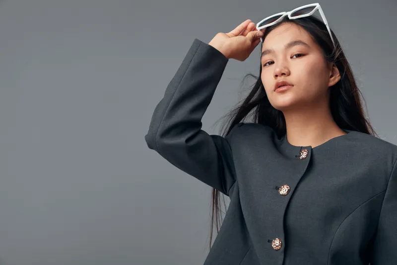 Young woman with long dark hair wearing a dark gray jacket adjusts white sunglasses on head against plain background. Portrait of confident female fashion model with natural makeup.