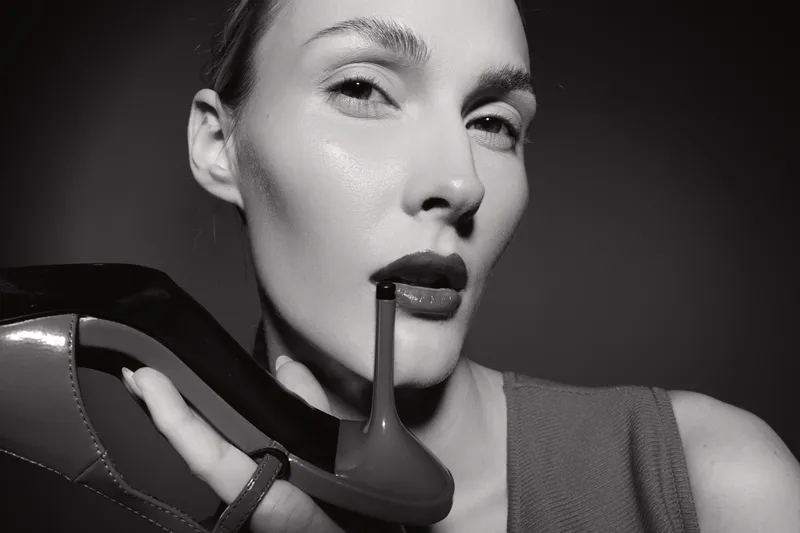 Close-up of a fair skinned Caucasian woman holding a high heel shoe near her face, with bold makeup and intense gaze in monochrome studio lighting.