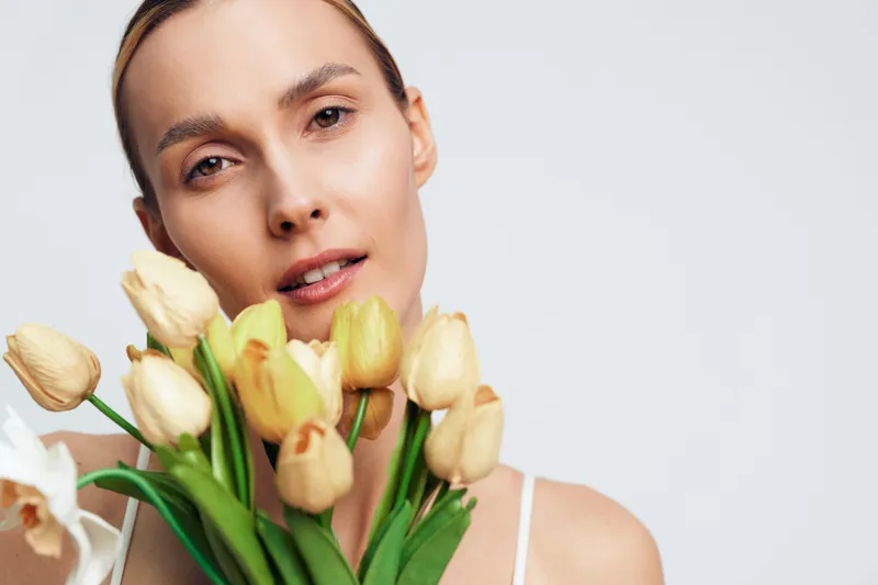 A young woman with natural skin holds a bouquet of yellow tulips close to her face against a plain light background. Concept for beauty, spring, and floral freshness.
