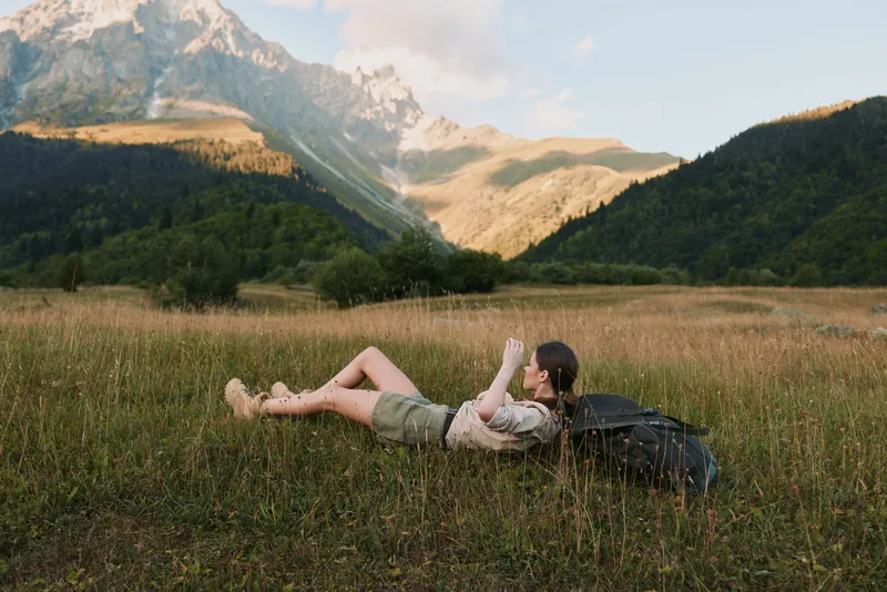 Young woman lying on grass in mountain valley reading book during sunny day. Concept of lifestyle, relaxation, and outdoor leisure in nature with backpack nearby.