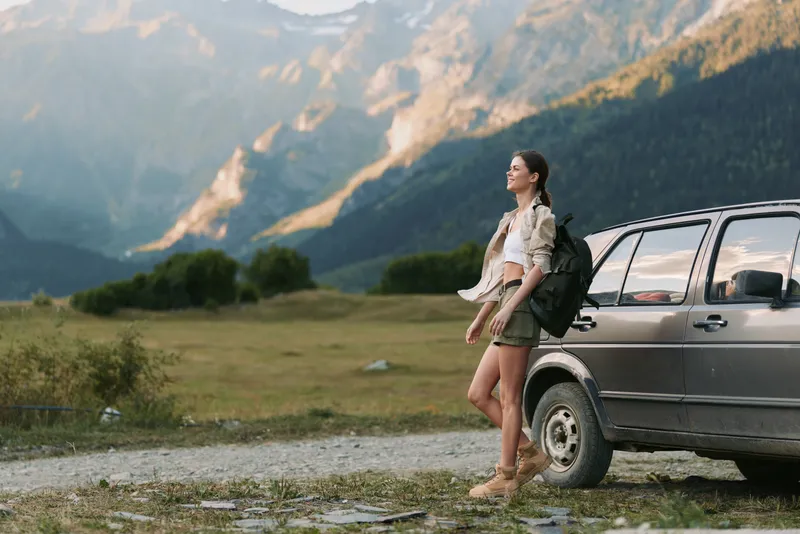 Young woman traveler standing near car in mountain landscape, enjoying nature, casual outfit, backpack, sunny day, relaxed mood, outdoor adventure lifestyle
