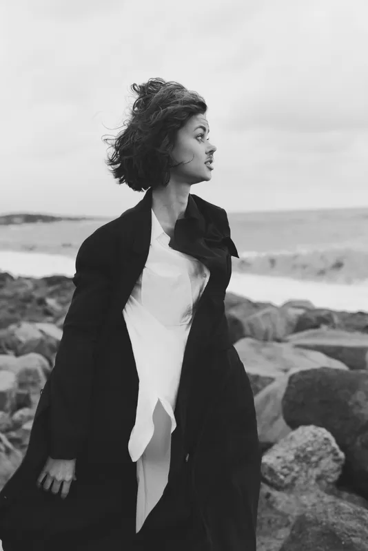 Woman in a long coat stands on a rocky shoreline, wind in hair, monochrome scene with sea in the distance, fashion photography vibe, moody and cinematic, capturing resilience and grace against the