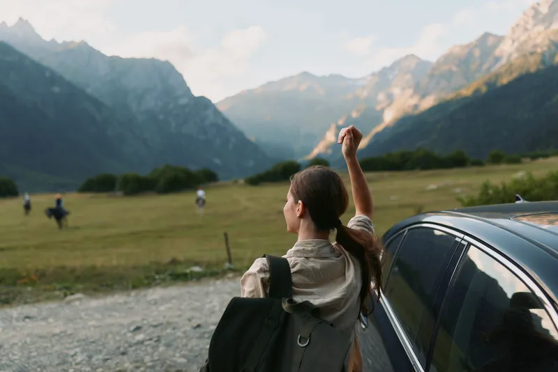 Young woman with backpack stands beside a car at the foothills, raising her arm in celebration against towering mountains under a clear blue sky, capturing a spontaneous travel moment.