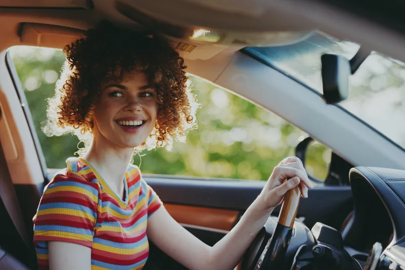 Young woman with curly hair smiles while driving a colorful striped tee in a sunlit car, exuding joyful, cinematic mood.