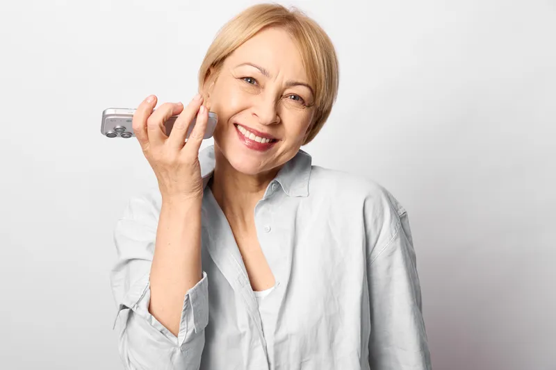 Older woman smiling in studio portrait, holding glasses and eyewear near face, relaxed hand gesture, gentle expression, soft white background, natural light, calm mood