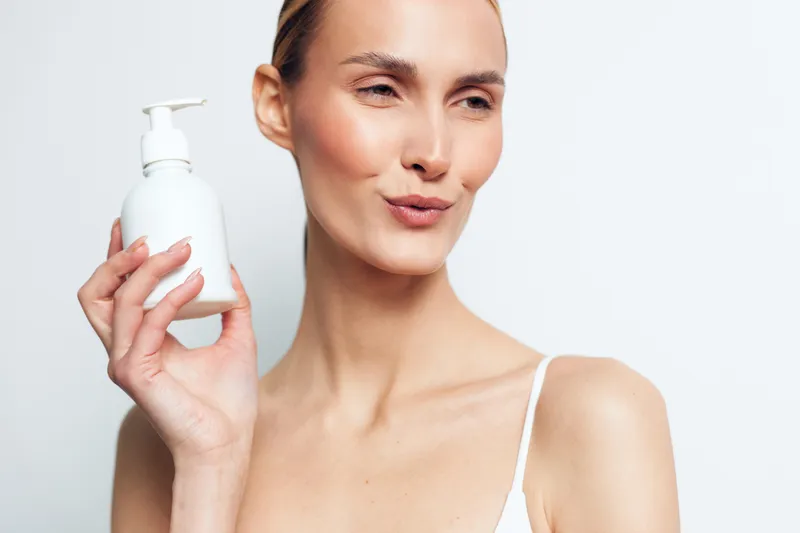 Young woman with clear skin holding white pump bottle near face on white background. Concept of skincare, beauty routine, hydration, and self-care with soft natural lighting.
