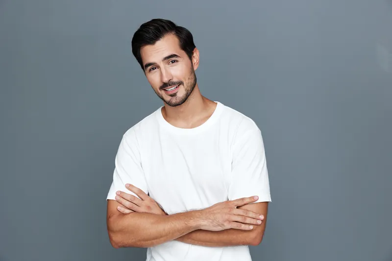 Full body smiling young man with dark hair stand confidently, wearing a plain white t-shirt, arms crossed, against a minimalist gray background. People lifestyle concept.