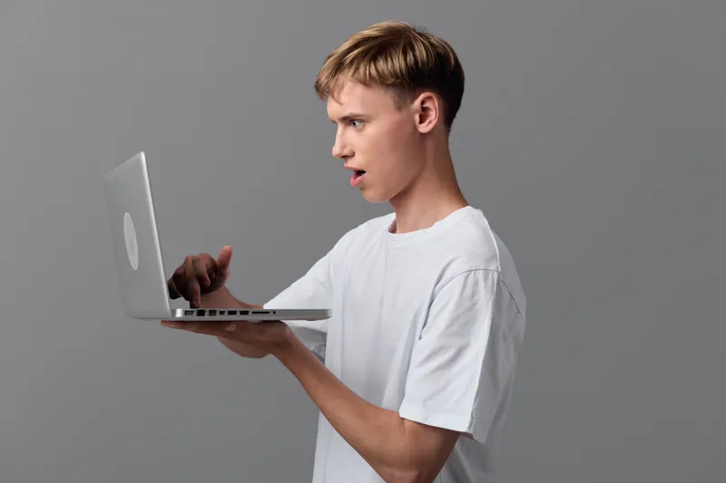 Surprised man holds a laptop aloft, exploring technology and digital learning with a casual vibe, white tee and neutral backdrop convey curiosity, innovation and hands on computing