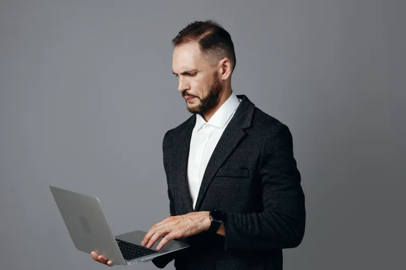 A focused professional man in a dark suit uses a laptop against an isolated gray background, conveying confidence, concentration, and business readiness.