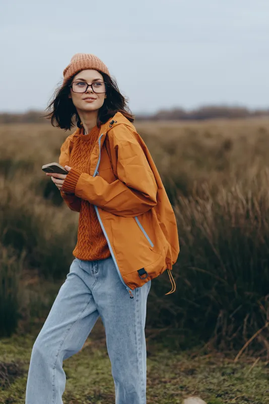 Woman outdoor lifestyle portrait, autumn field background, orange jacket, knit hat, glasses, warm scarf, standing on path with gentle smile, natural daylight, casual travel.