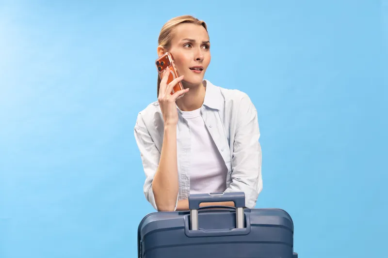 Young woman in casual clothes holding phone to ear and sitting behind blue suitcase on blue background. Concept of travel, communication, and waiting at airport or station.
