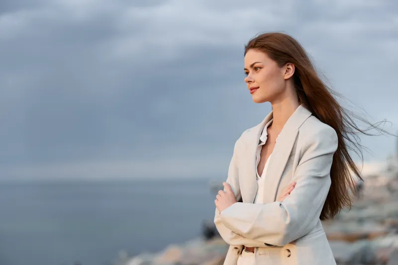 Woman in beige coat standing confidently outdoors with arms crossed, natural makeup, long hair flowing, overcast sky and rocky beach background, fashion theme