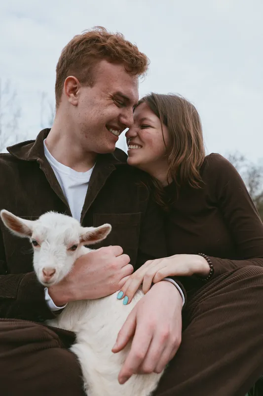 A loving couple outdoors cuddle a playful goat kid, sharing a tender moment in natural light. The scene conveys warmth, companionship, and simple rural happiness, highlighting lifestyle with animals