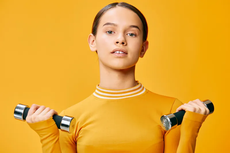Woman in a bright yellow top holds dumbbells for a focused fitness workout, in a studio background, showcasing strength training and athletic lifestyle.