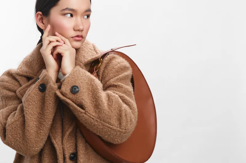 Asian woman dressed in a warm brown coat with a large bag stands thoughtfully against a neutral backdrop, projecting chic urban style, poise, and quiet confidence.