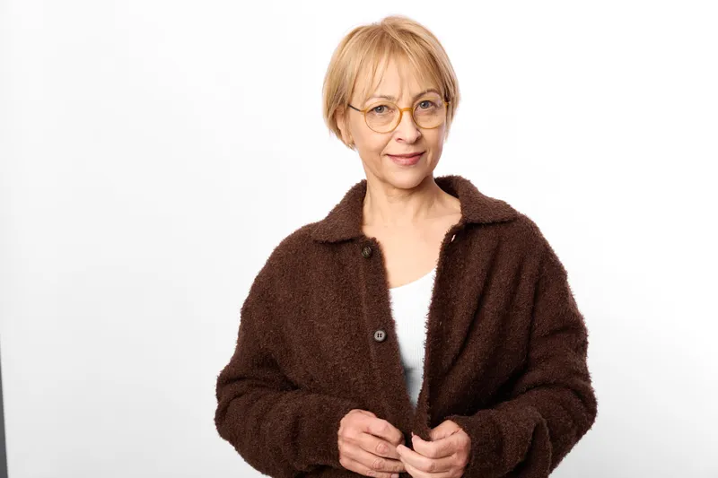 Older woman with glasses, brown coat portrait in studio background, calm smile, short blonde hair, hands near buttons, front view, soft even light