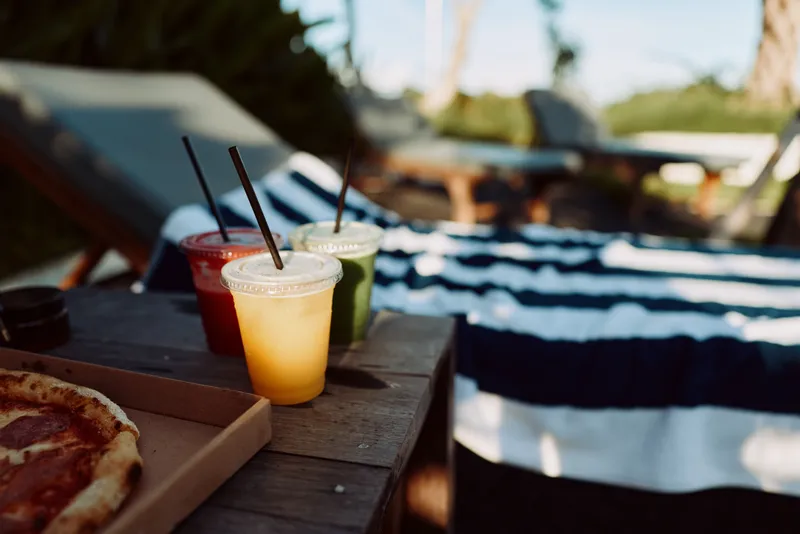 A poolside scene with colorful drinks and a pizza on a wooden table, sunlit loungers in the background. A lifestyle moment capturing casual summer leisure, refreshment, and social downtime.