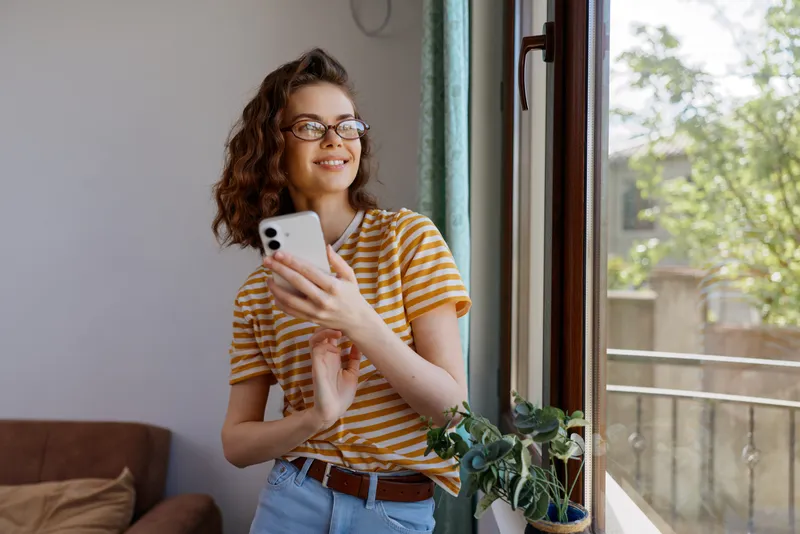 A smiling woman in a striped shirt stands by a sunlit window at home, holding a smartphone. A casual lifestyle moment highlighting everyday comfort and indoor brightness.